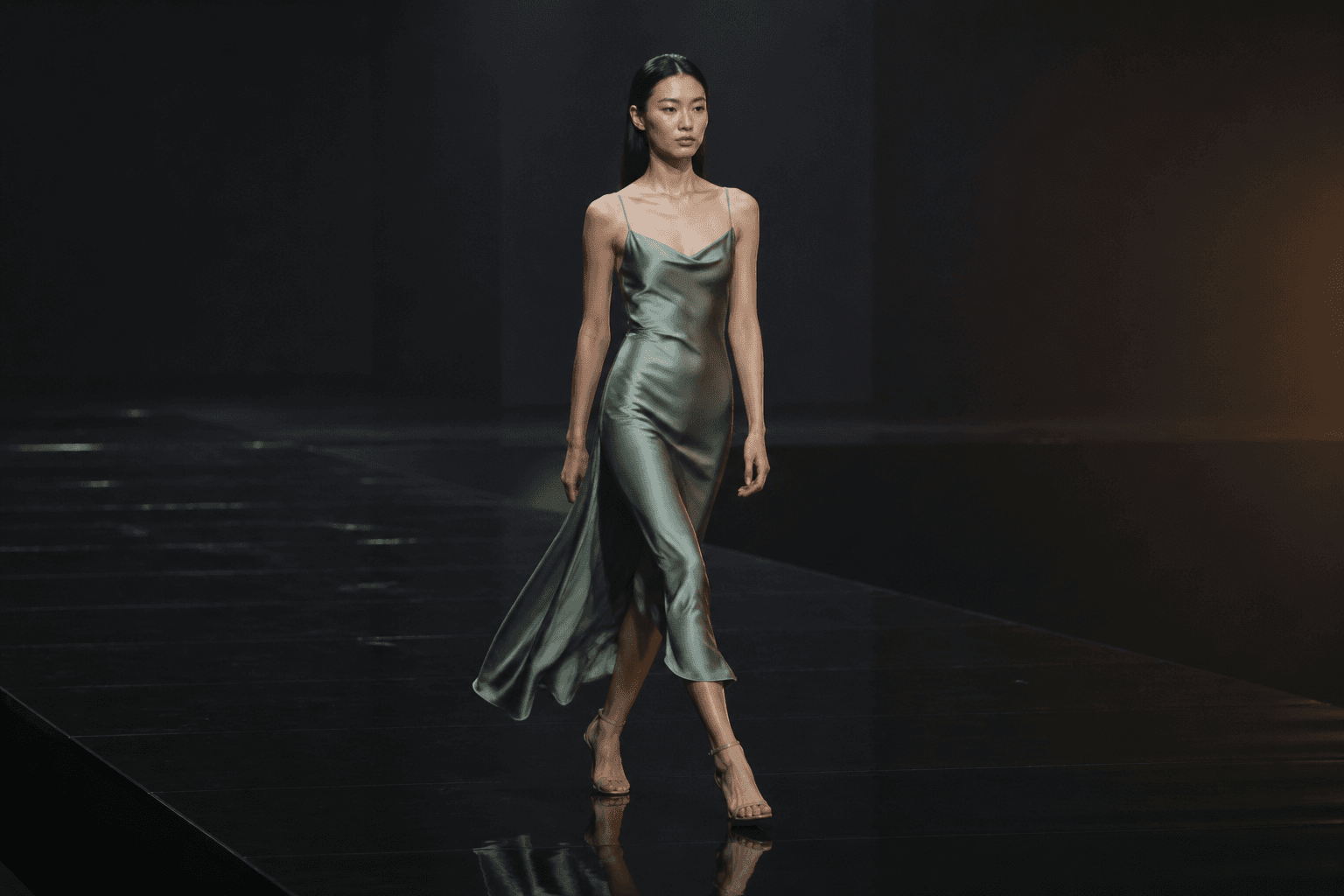 Editorial runway film still — model in sage silk slip on a matte black infinity stage.